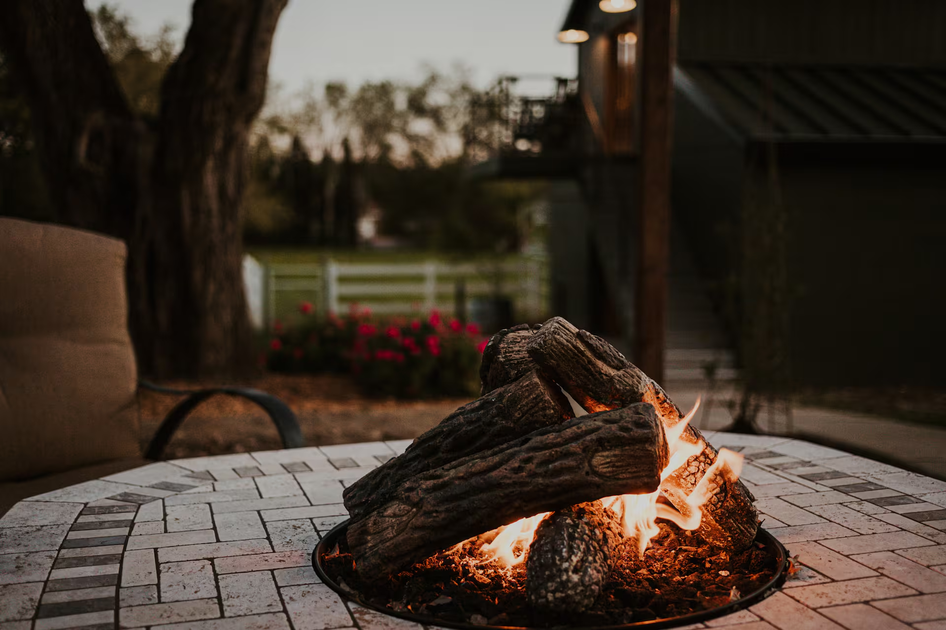 A circular brick-paver fire pit with live flames dancing over stacked logs at dusk, a cushioned patio chair to the left, pink garden flowers blooming behind a white fence, and the barn softly lit beyond