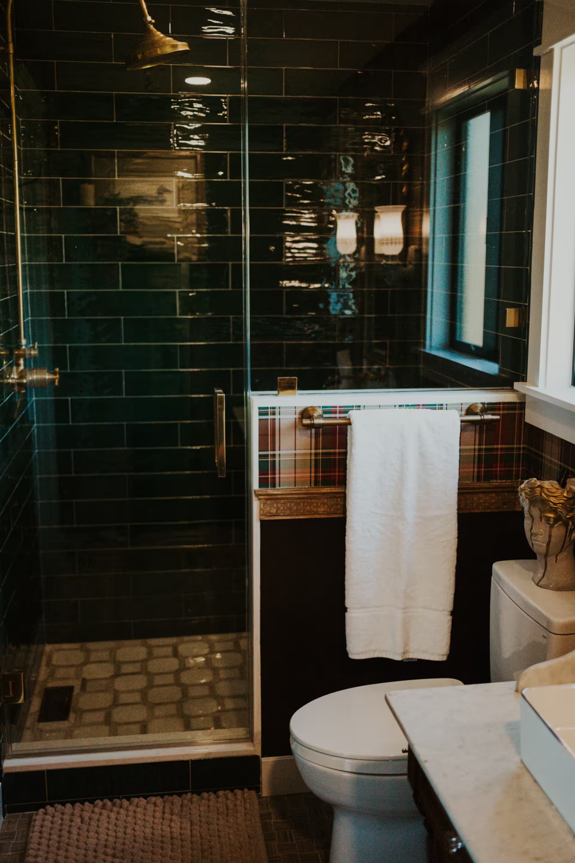 The ensuite bath with glossy deep-green subway tile, aged brass fittings, tartan plaid wallpaper, and a frameless glass walk-in shower